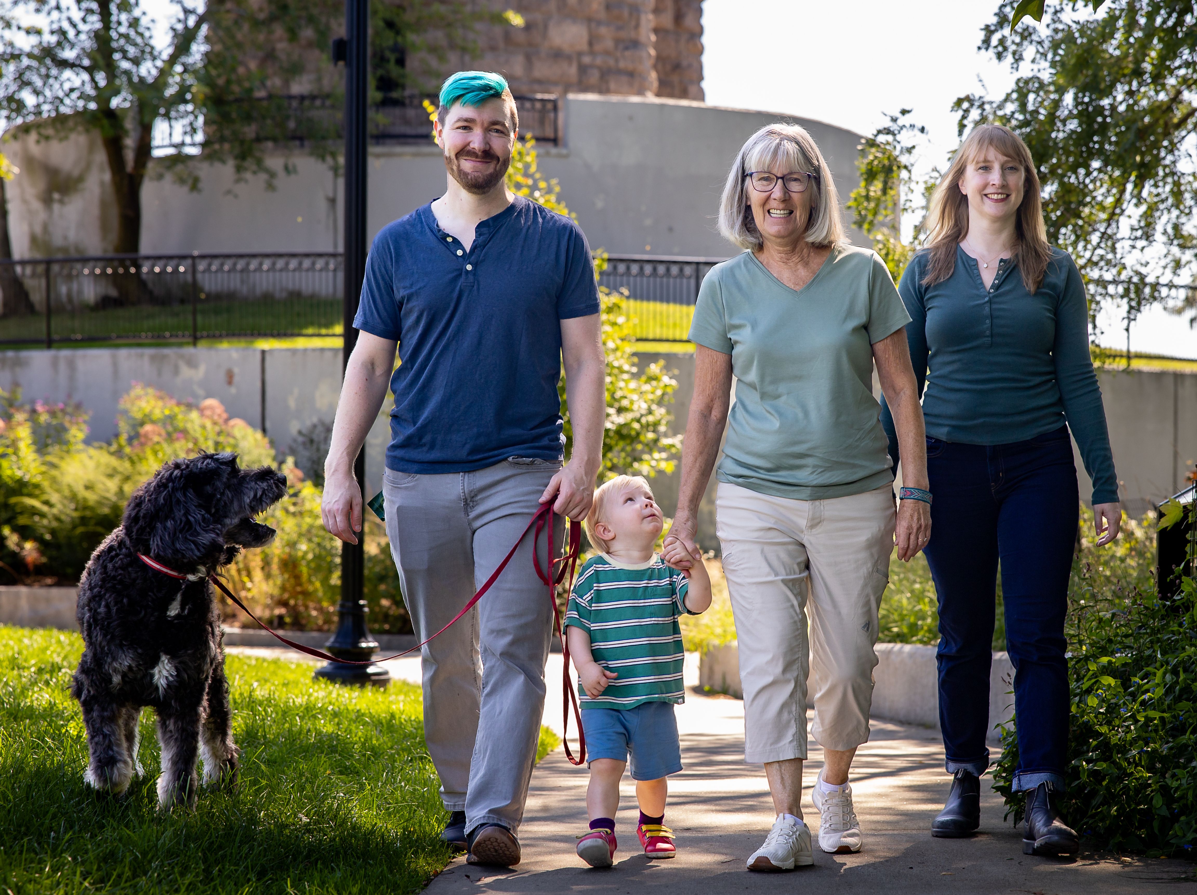 Derrick walking in a park with his wife, toddler son, mother, and dog. Prospect Hill tower is in the background.